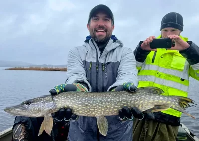 A smiling fisherman showing off his catch while his friends takes a photo of him and his fish