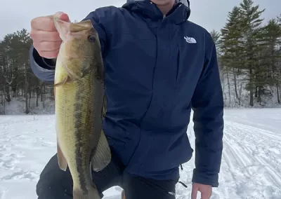 A young man dressed in winter gear, kneeling on an icy lake, holding a large mouth bass he caught ice fishing