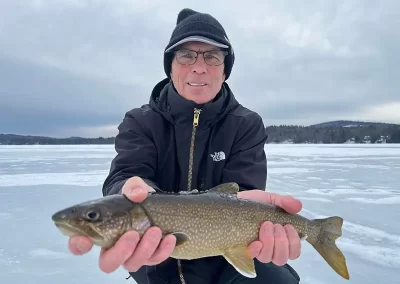 An older gentleman dressed in winter gear holding a fish on a clouding day on the icy lake