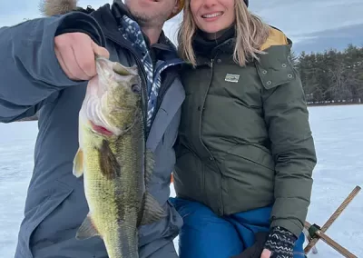 A proud couple kneeling on the icy lake on a cloudy day where the man is holding up the fish they caught.