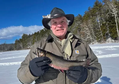 An older many in a sunny wintery scene holding a fish he caught ice fishing