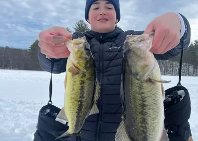 A boy in wintery gear on an icy lake holding two large mouth bass he caught while gloves dangle by his side