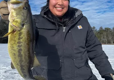 A smiling woman in winter gear with a wintery background, holding the large mouth bass she caught while ice fishing