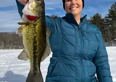 A smiling woman dressed in winter clothes, kneeling in the snow, smiling and holding a fish she caught