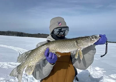 A person dressed in winter gear from head to toe on a wintery, icy lake holding a huge Northern Pike